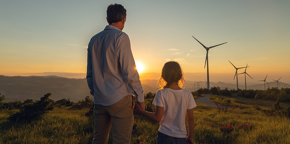 A man and a small child look out at a windmill at sunset.