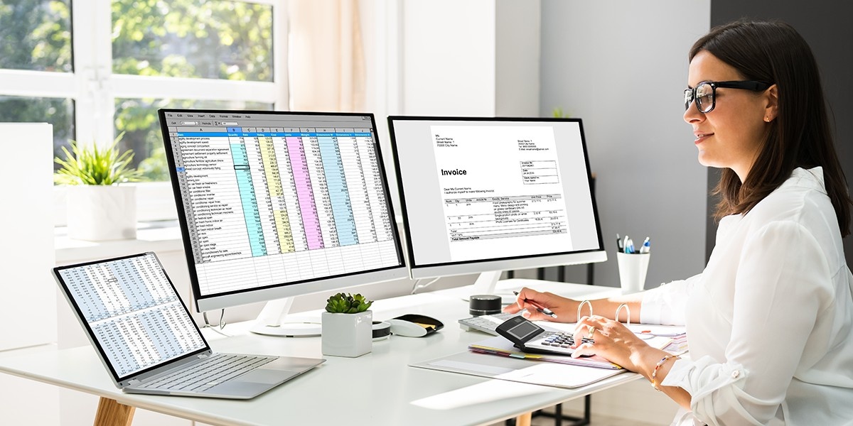 A woman sits at a desk with two computer monitors and a laptop.