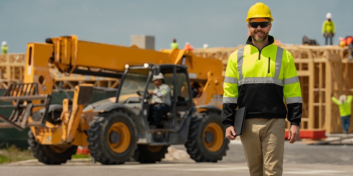 A worker in a high-vis jacket and hard hat walks away from an excavator.