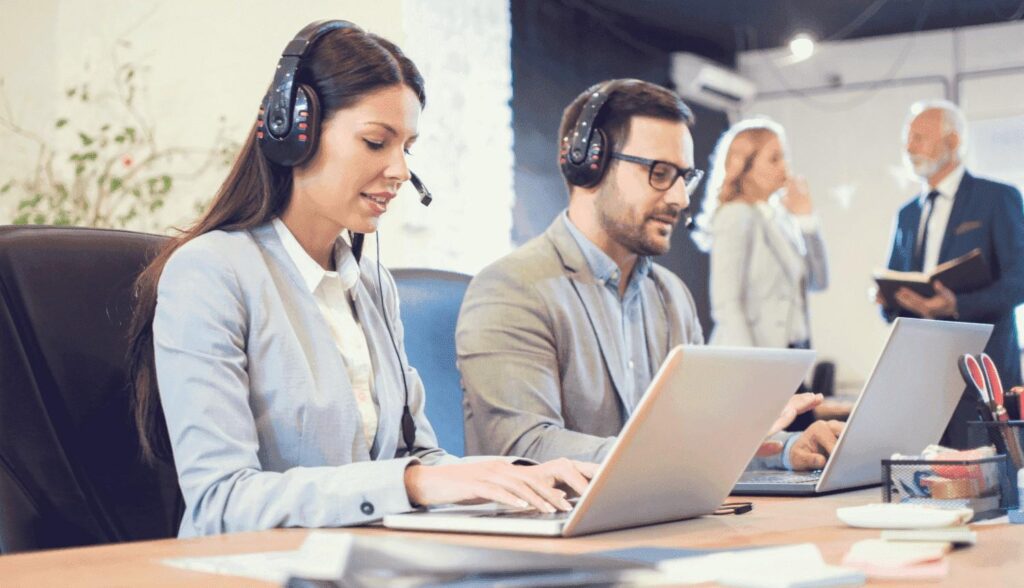 Two customer service agents sit at a desk with headsets on.