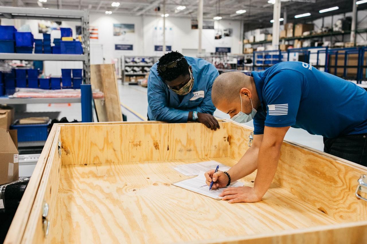 Two workers stand over a wooden desk writing notes on a sheet of paper.