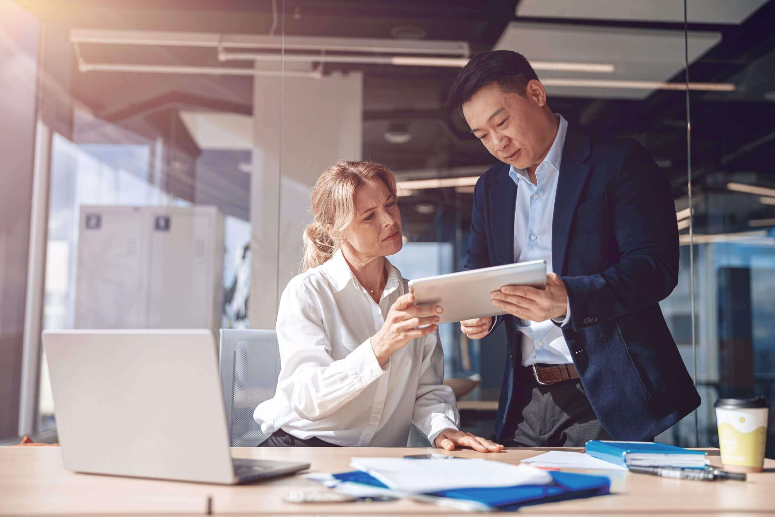 A man and a woman in business attire look at a document together at the woman's desk.