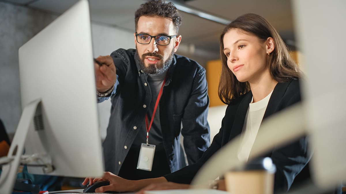 A woman works on her computer while a man stands by her desk pointing at the computer screen.