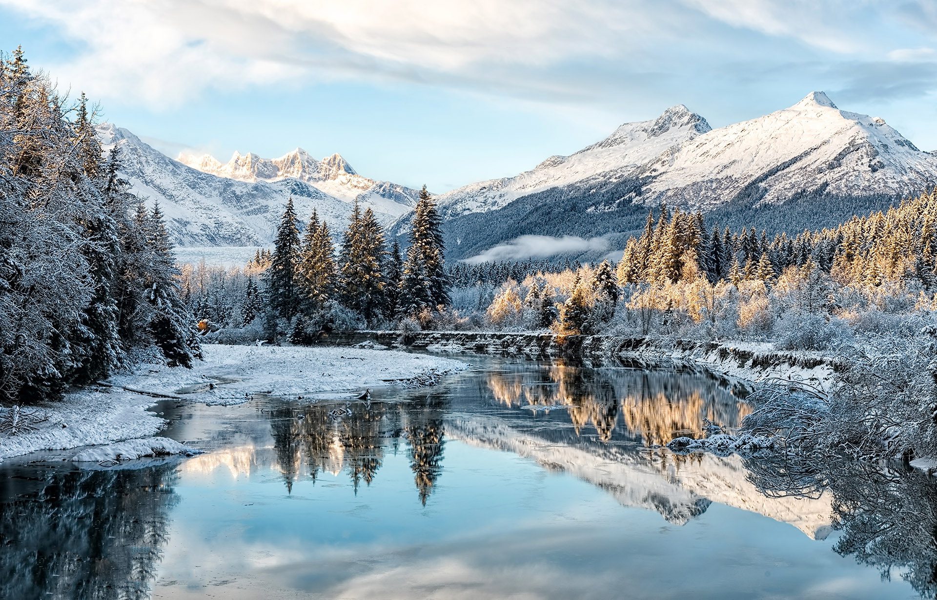 A wintery forest landscape with a lake in the foreground and snowy mountains in the background.