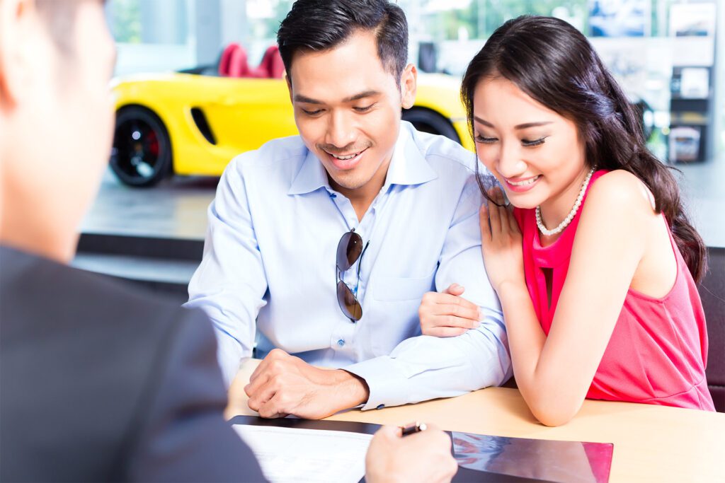 An Asian couple smiles while signing a contact with a yellow convertible car in the background.