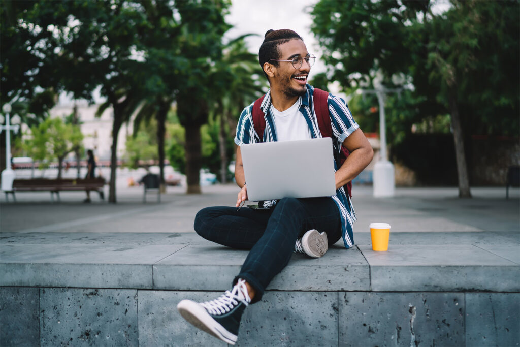 A young African-American man sits on a bench in a tropical campus area, smiling and working on a laptop.