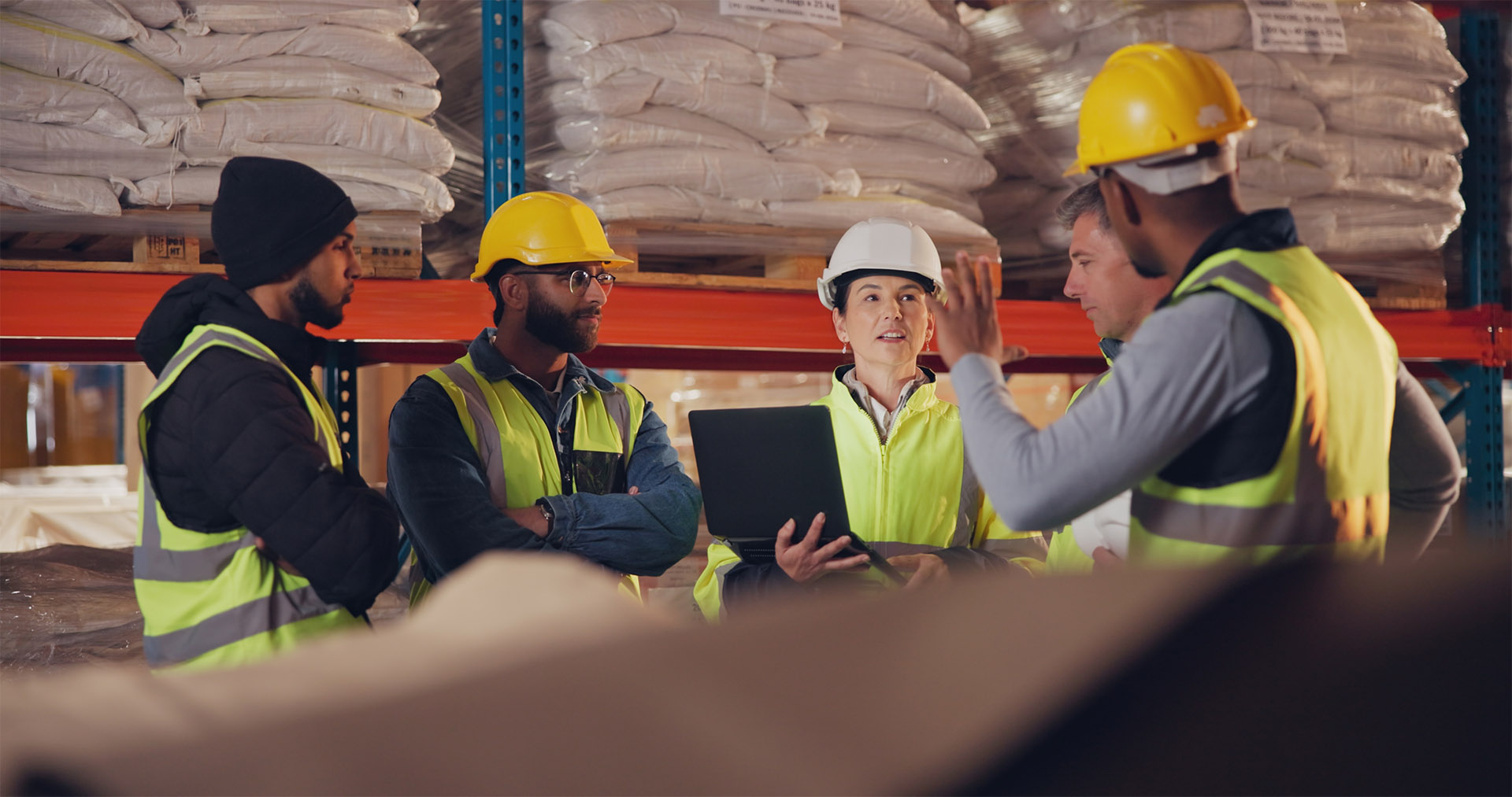 A group of workers in high-vis jackets and hard hats stand in a warehouse discussing processes.