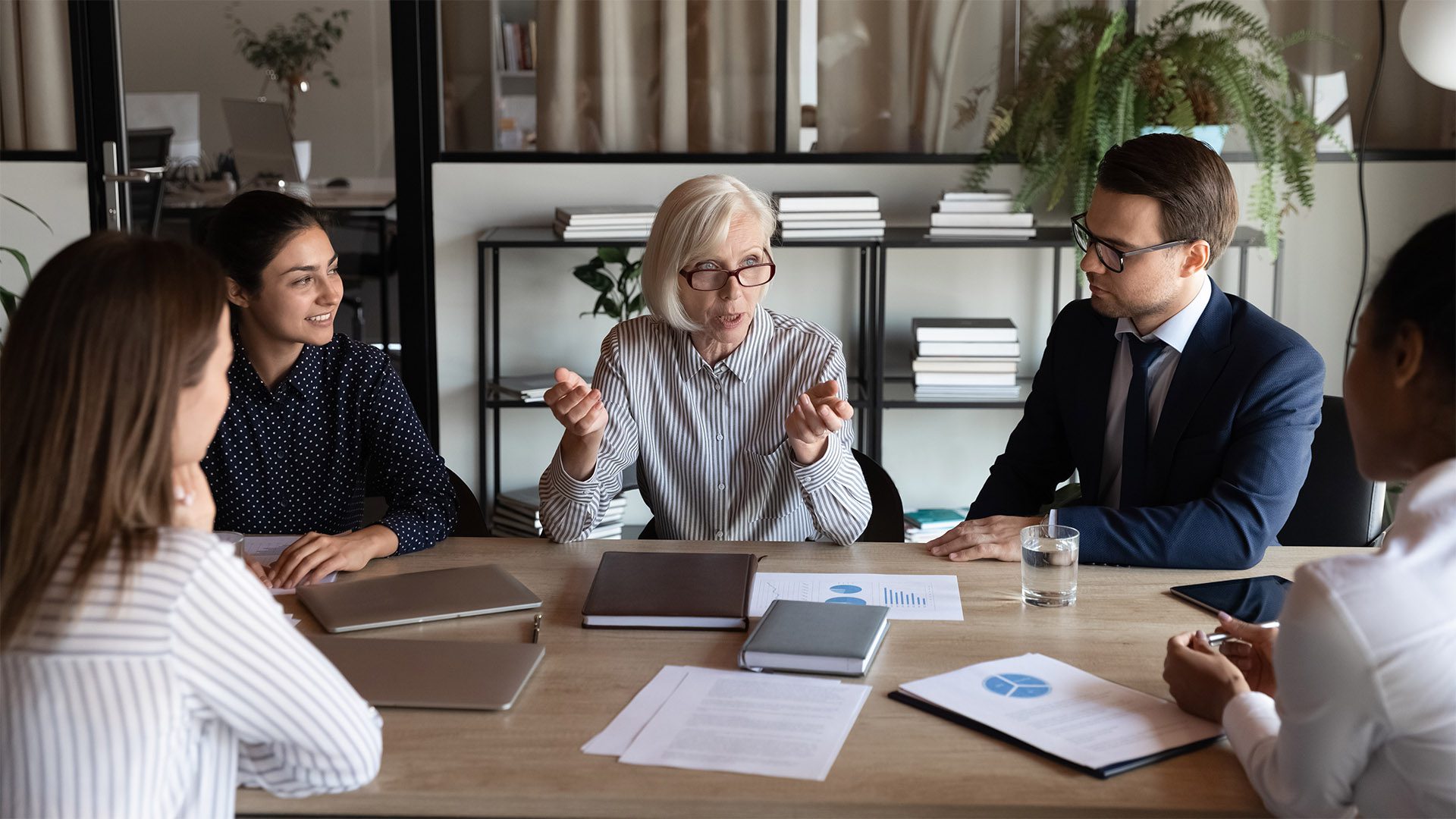 An older woman sits at a table discussing with her team, who are gathered around the table.