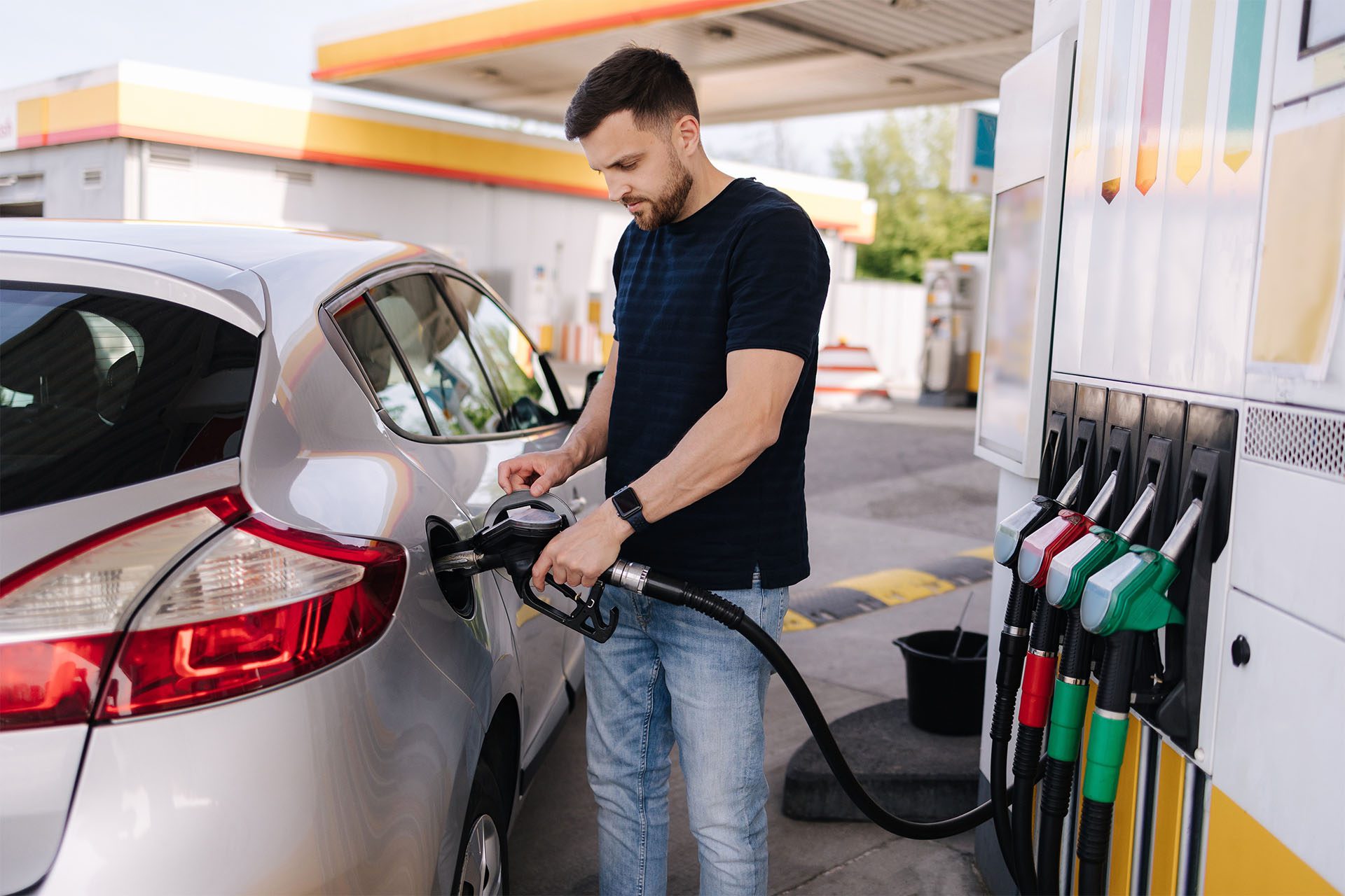 A young man in a black t-shirt and jeans refuels his car at a gas station.