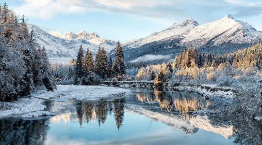 A wintery forest landscape with a lake in the foreground and snowy mountains in the background.