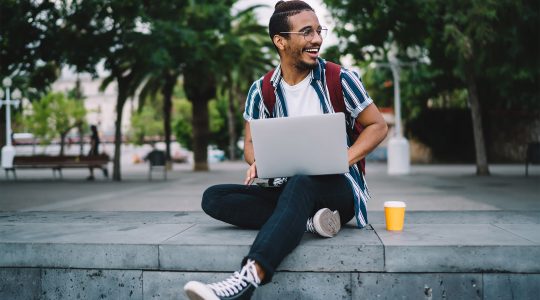 A young African-American man sits on a bench in a tropical campus area, smiling and working on a laptop.