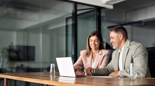 Two middle-aged businesspeople sit at a desk and participate in a video call on a laptop.