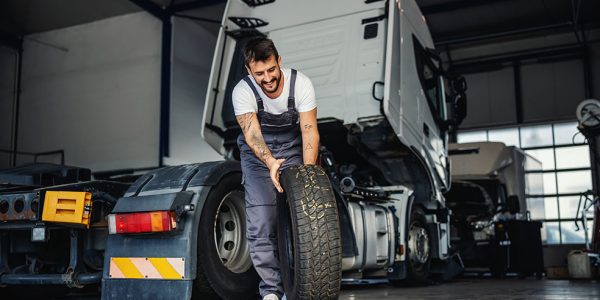 A mechanic in overalls rolls a tire away from the cab of an 18-wheeler.