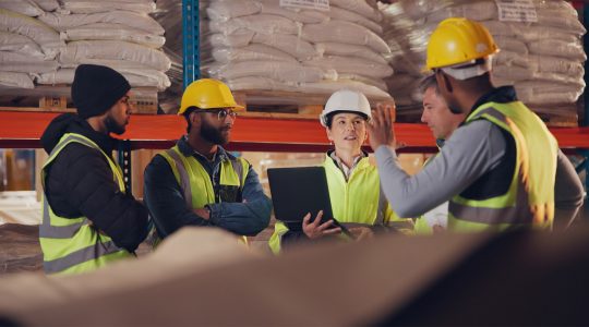 A group of workers in high-vis jackets and hard hats stand in a warehouse discussing processes.