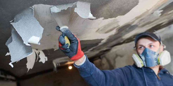 A contractor in protective equipment removes spackle from a wall.